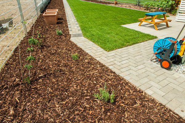 A fresh hardwood mulch bed lining a backyard walkway, with a manicured lawn and patio in view — same-trip delivery and spread.