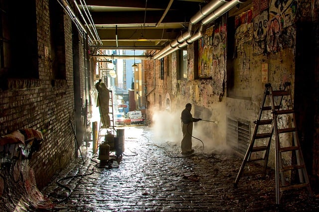 Two workers repair and clean an alley. One is on a ladder repairing the left wall, and the other is power washing the right wall.